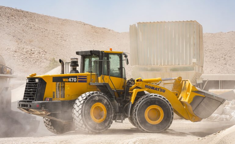 A yellow construction vehicle drives through a dirt field, kicking up dust as it moves across the terrain.